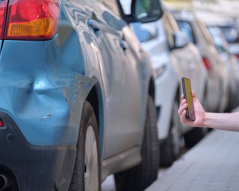 A hand holding a phone beside a dented car parked on the street