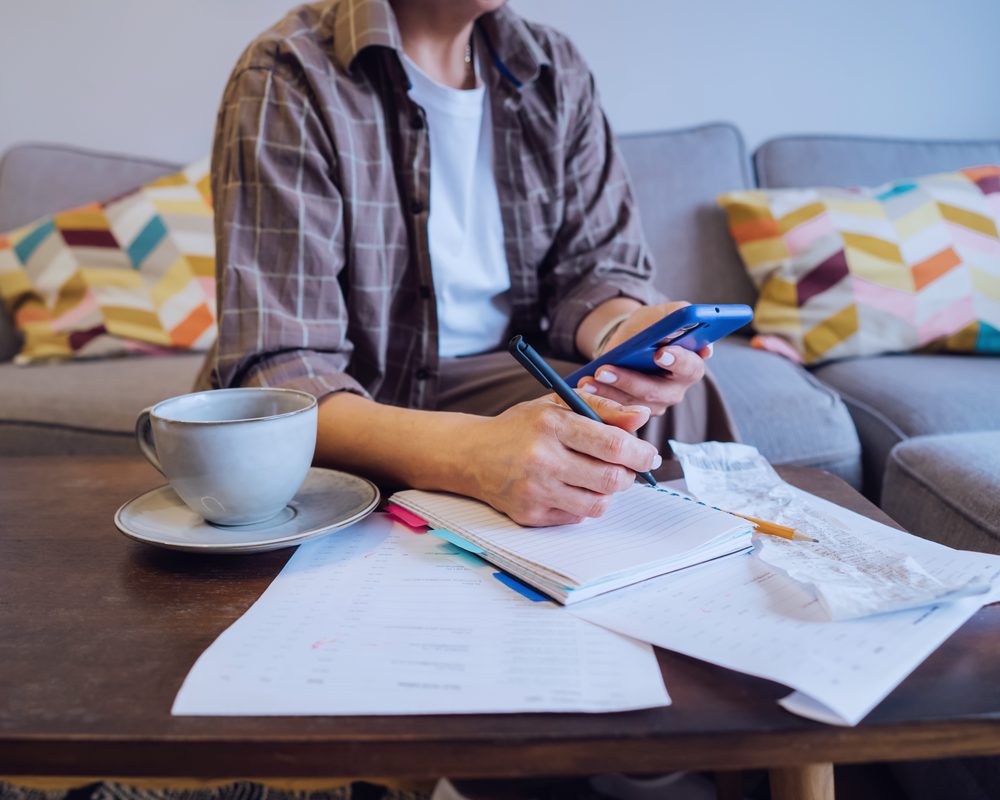 A person at home with a notebook, phone and coffee, going through paperwork